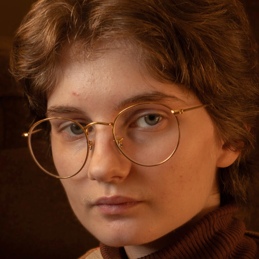 A photographic portrait of a young adult wearing large gold rimmed glasses.